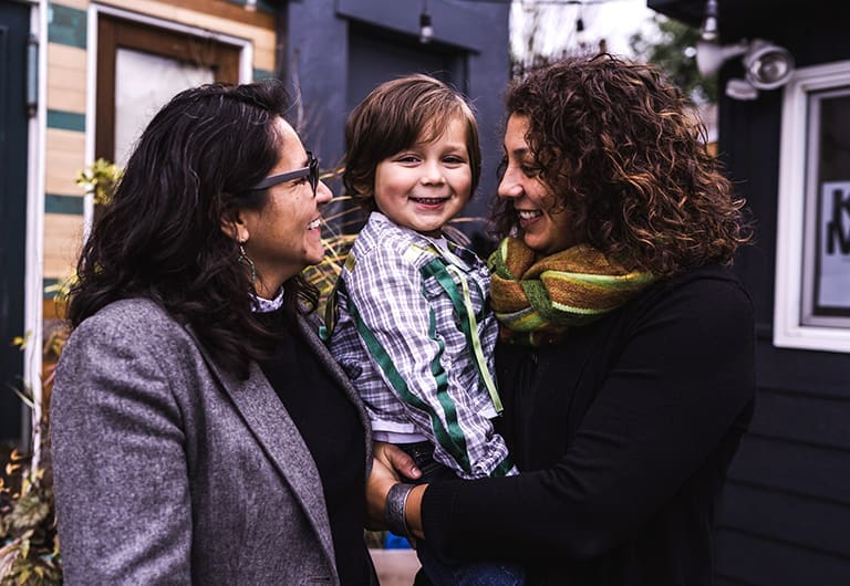Two women smiling and looking inwards towards a child that one of them is holding. Together they make a beautiful family.