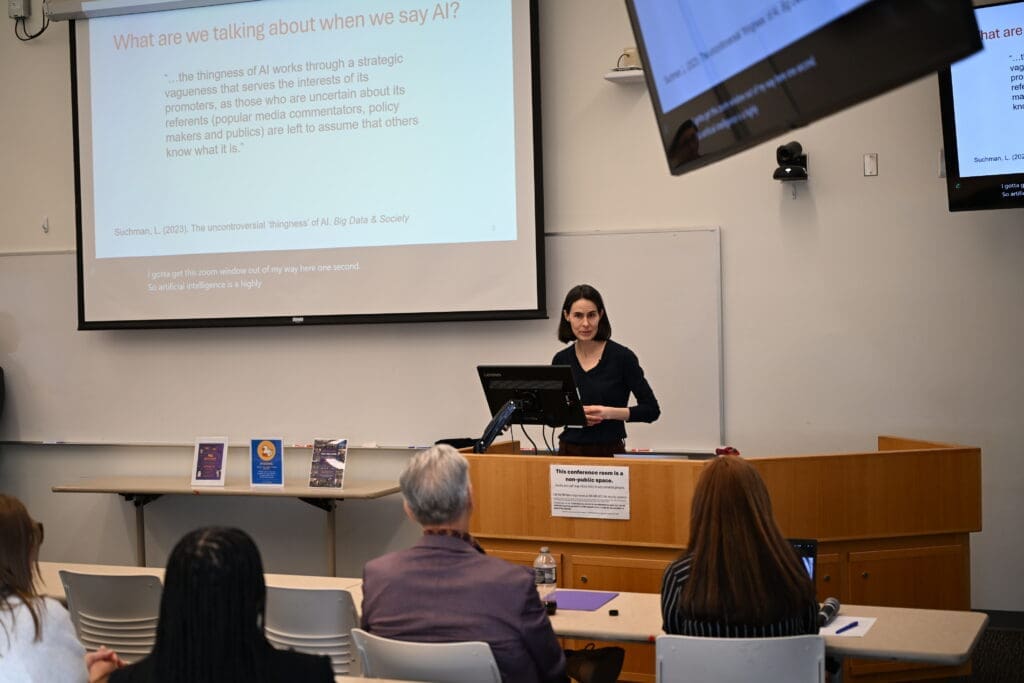 Clara Berridge gives a research talk from a podium in Social Work Room 305A. She is wearing a black sweater. To her right is a slideshow on a projector and in the forground, her colleagues look on.