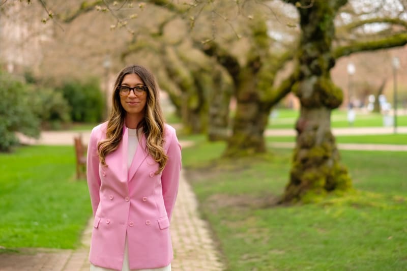 Hannah Scheuer stands in front of cherry blossom trees on the UW campus. She is wearing a pink blazer and glasses.