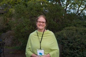 J'May Rivara stands in front of a large shrub. She is wearing a green top and smiling.