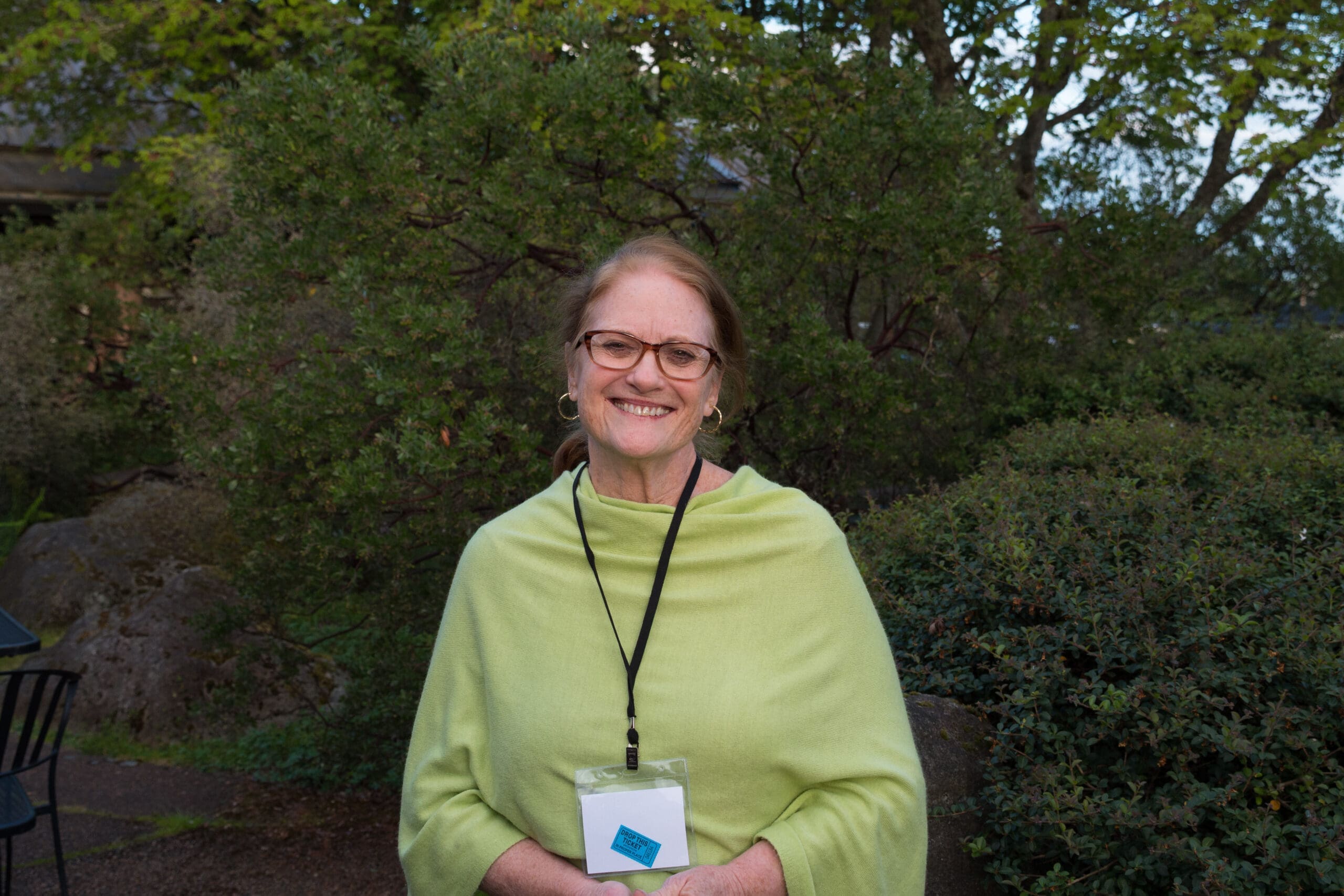 J'May Rivara stands in front of a large shrub. She is wearing a green top and smiling.