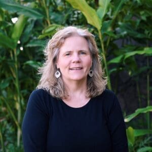 Professional headshot of Karen Fredriksen Goldsen, PhD. Photo features an older white woman with blonde hair and a black t-shirt standing outside in front of a tall green bush.