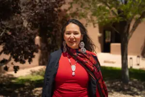 Professor Johnson-Jennings smiles on a sunny day, while wearing a red and black scarf over a red shirt, and large, circular earrings.