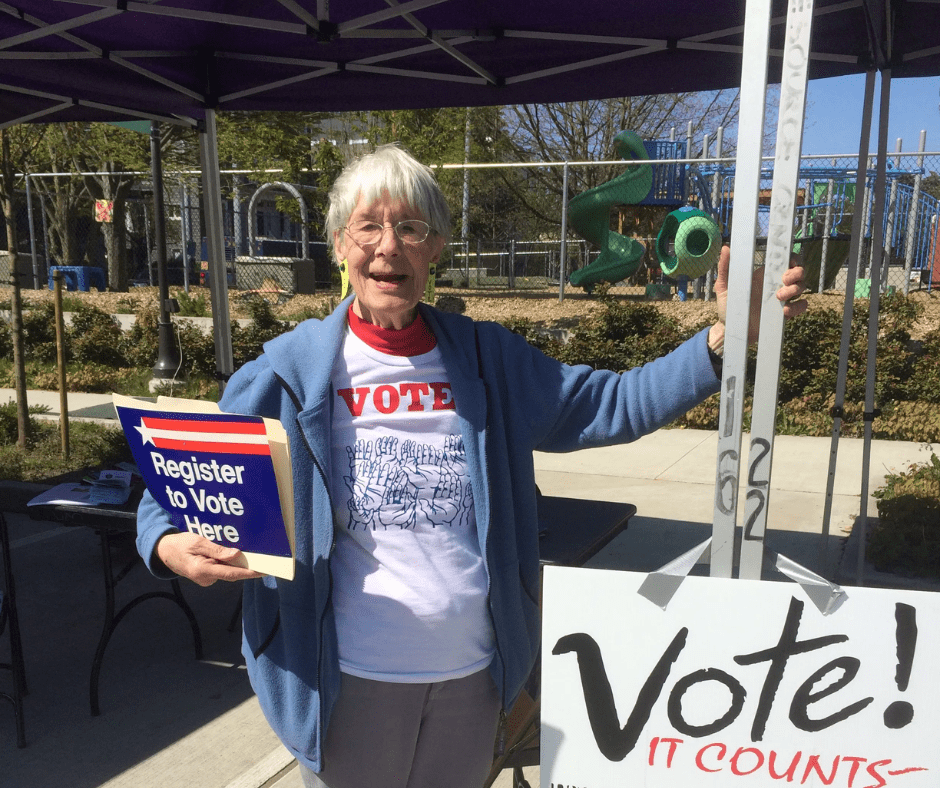 Nancy Amidei is smiling and standing next to a sign that says "Vote, it counts!" She is wearing a blue jacket, white shirt, and gray pants.