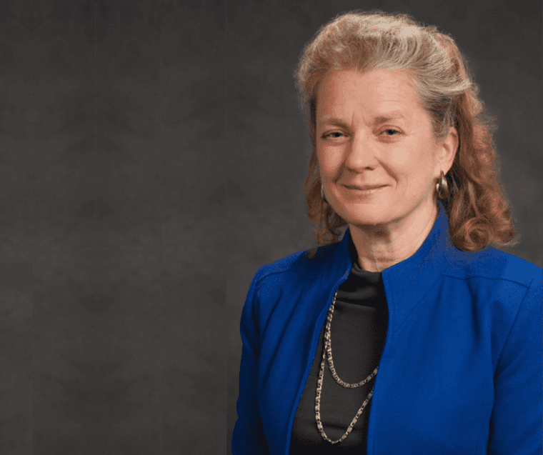 Paula Nurius wearing a blue blazer in front of a grey background.