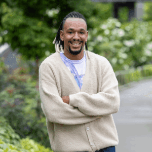 Isaac folds his arms and smiles into camera. They are wearing a cream-colored sweater, and standing outside in a walkway with greenery behind them.