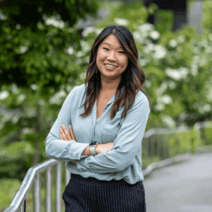 A young woman stands, arms folded and smiling, in an outdoor walkway. Greenery and white flowers surround her.