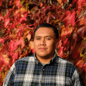 Headshot of Matt Frank. He's wearing a navy-blue and white flannel while standing in front of a wall covered in red leaves.