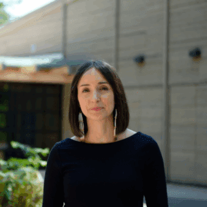 A young dark-haired woman wearing a black sweater looks into camera. She's in front of a building on a sunny day.