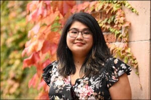 Silver Lee smiles for the camera while wearing a black and pink floral top. Red leaves hang from the wall in the background.