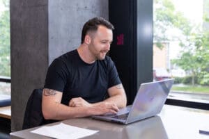 A male student wearing a black shirt sits at a table, using a laptop, in a dormitory common area.