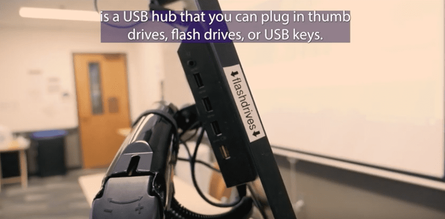 Side view of a desktop monitor showing multiple USB ports. The monitor is in a classroom with a whiteboard behind it.