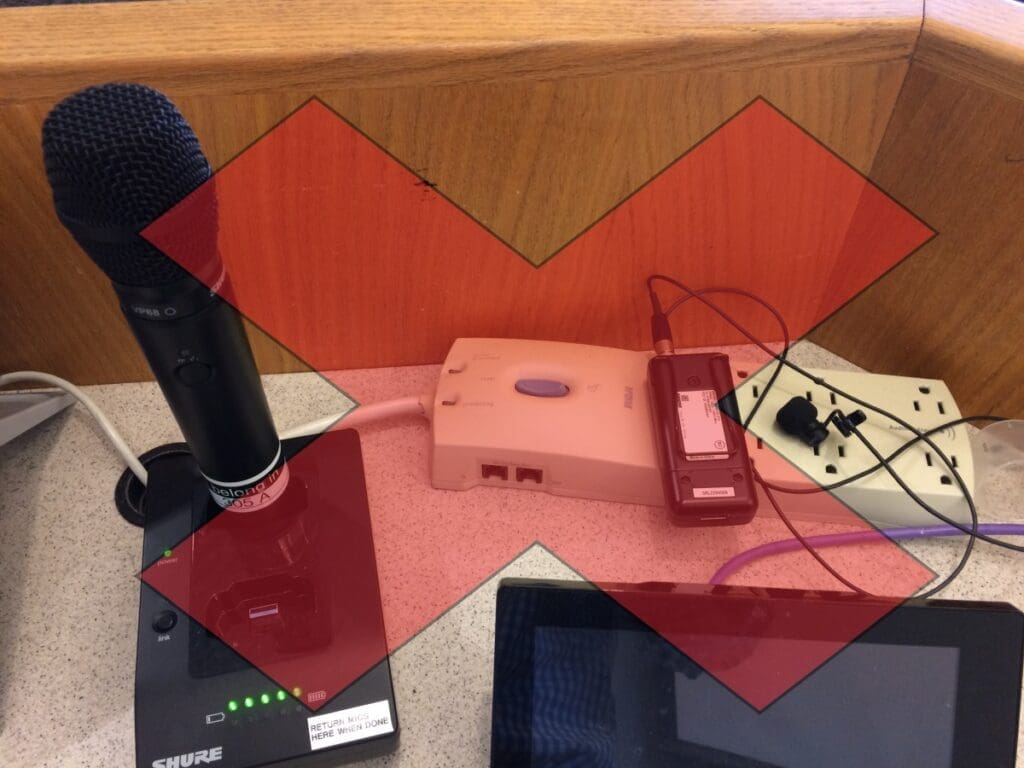 Black microphone, charger, touchscreen display and cords sitting on a podium with a large red X overlaying the entire image.
