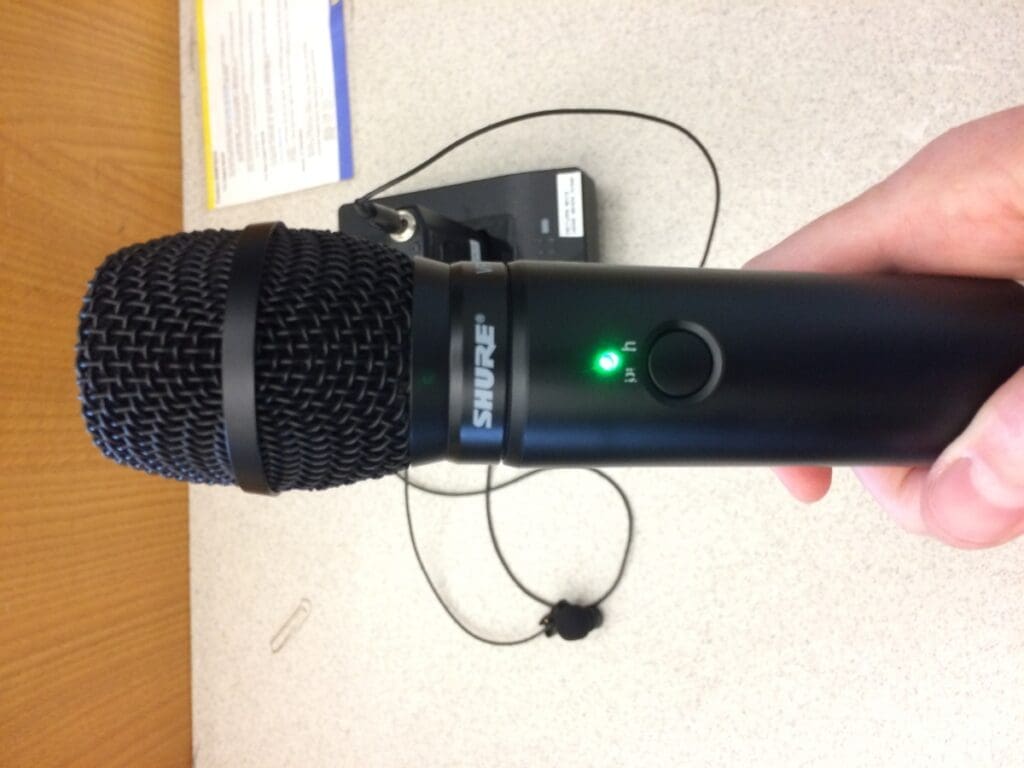 Black microphone, with a green light on in the middle, being held up by a white skintone hand.